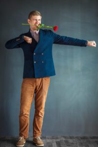 Handsome young man dancing with a red rose in his mouth against grey wall background
